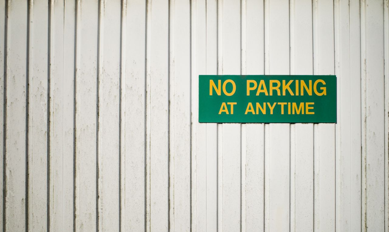 A green 'No Parking at Anytime' sign on a white weathered wooden wall.